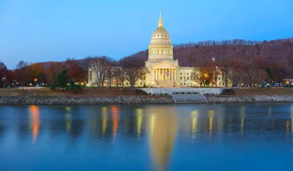 A grand, illuminated capitol building at twilight reflects elegantly on the calm waters of a nearby river.