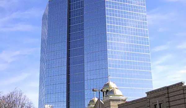 A tall glass skyscraper overlooks smaller traditional buildings under a clear blue sky.