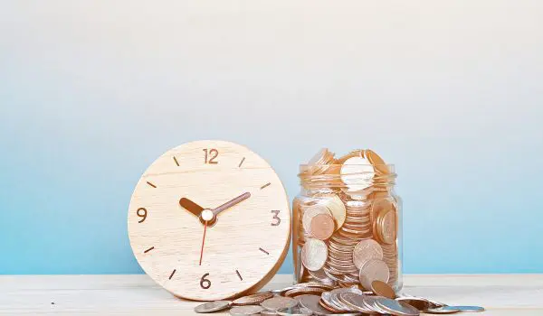 A wooden clock is placed next to a jar filled with coins, with more coins scattered around the jar.