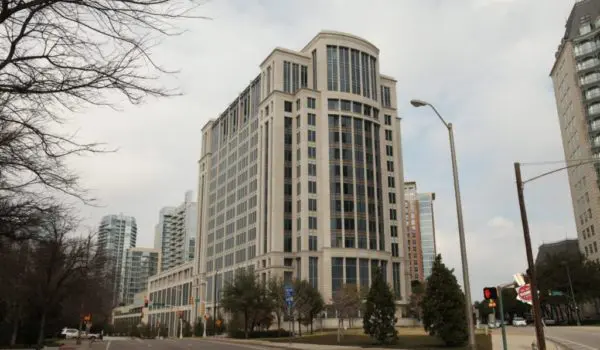 A wide street is lined with tall, modern buildings and trees, leading to a large corner building with a rounded design element on top.