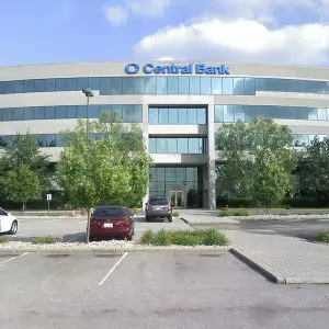 A modern multi-story building with a "Central Bank" sign, surrounded by a parking lot with several parked cars and trees.
