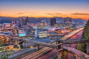 A vibrant cityscape is captured at dusk, featuring illuminated buildings and intersecting highways against a backdrop of rolling hills and a colorful sky.