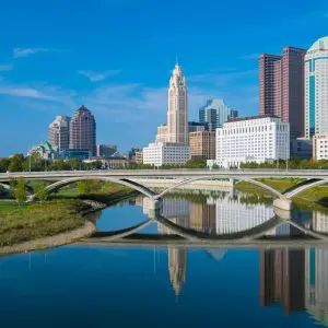 A city skyline with modern buildings is reflected in a calm river under a blue sky.
