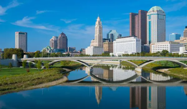 A city skyline with modern buildings is reflected in a calm river under a blue sky.