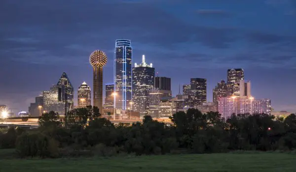 A city skyline illuminated at dusk with a mix of modern skyscrapers and a distinctive spherical structure, set against a twilight sky with greenery in the foreground.