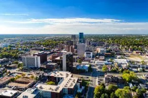 An aerial view of a downtown district showcases a mix of high-rise buildings, urban streets, and scattered greenery under a blue sky with light cloud cover.