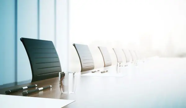 A row of black chairs alongside a modern conference table with a glass of water, indicating a professional or business setting, with bright natural light in the background.
