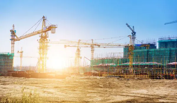 A construction site with multiple cranes and a scaffolding structure illuminated by bright sunlight.