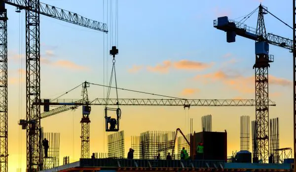 Silhouetted construction cranes tower over a building site against a sunset sky.
