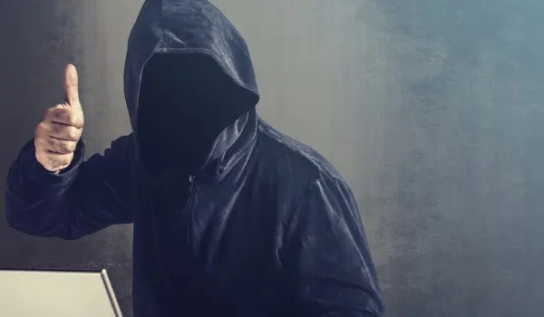 A hooded individual gives a thumbs-up gesture in front of a laptop on a desk against a textured backdrop.