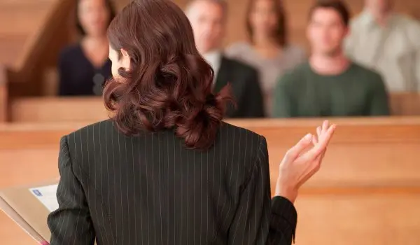 A person with dark hair is standing facing an audience, gesturing with their right hand, possibly speaking, while holding a tablet in their left hand.
