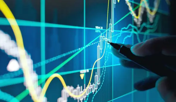 A hand holding a pen points to a luminous stock market graph on a digital screen, suggesting a focus on financial analysis or trading activities.