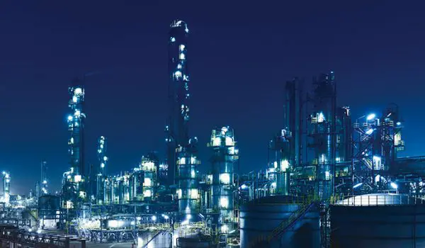 A nighttime scene of an illuminated industrial complex with multiple structures and bright lights set against a dark blue sky.