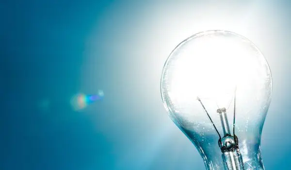 A clear light bulb is illuminated against a bright blue background with light flares.