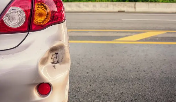 A silver car with a dented bumper is parked beside a road with a yellow dividing line.