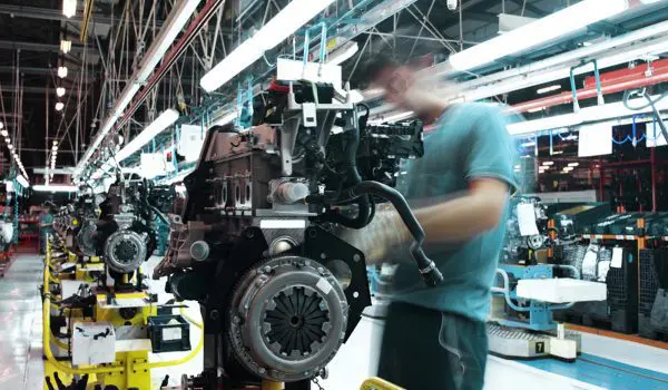 A worker assembles car engines on a factory production line with industrial equipment and fluorescent lighting overhead.