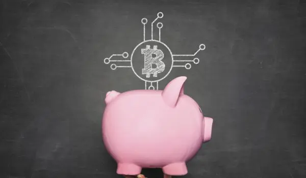 A pink piggy bank is being held up against a chalkboard background with a Bitcoin and circuit diagram drawn above it.