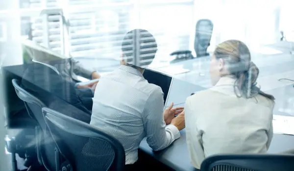 Two professionals are engaged in work at their computers in a modern office setting, with a view through a glass partition.
