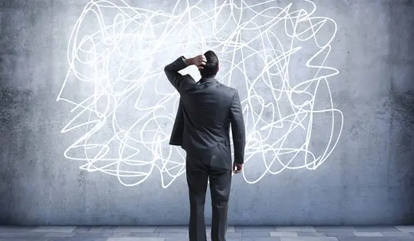 A person in a suit stands confused in front of a chaotic, scribbled mess on a wall.