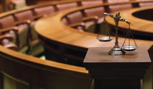 A set of balanced scales sits on a pedestal in an empty courtroom.