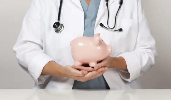 A healthcare professional in a lab coat with a stethoscope around their neck is holding a pink piggy bank.