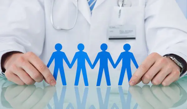A person wearing a white lab coat and a stethoscope holds paper cutouts of blue human figures linking hands on a reflective surface.