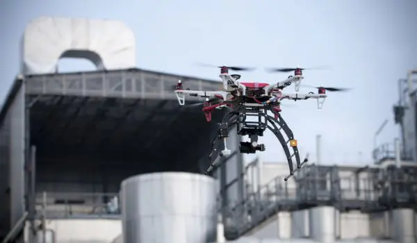 A drone equipped with electronic components is flying in front of industrial silos.