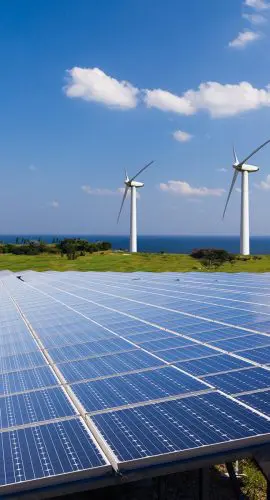 A solar panel array in the foreground with wind turbines in the background under a clear blue sky.