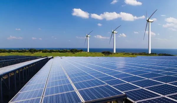 A solar panel array in the foreground with wind turbines in the background under a clear blue sky.