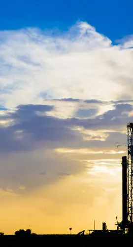 Industrial towers stand silhouetted against a vibrant sunrise or sunset with scattered clouds in the sky.