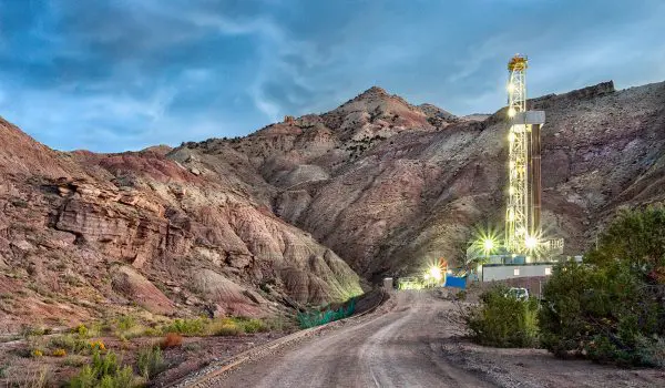 A lit drilling rig stands against a backdrop of rugged hills under a twilight sky.
