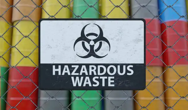 A weathered "Hazardous Waste" sign is attached to a chain-link fence, behind which various colored barrels are stored.