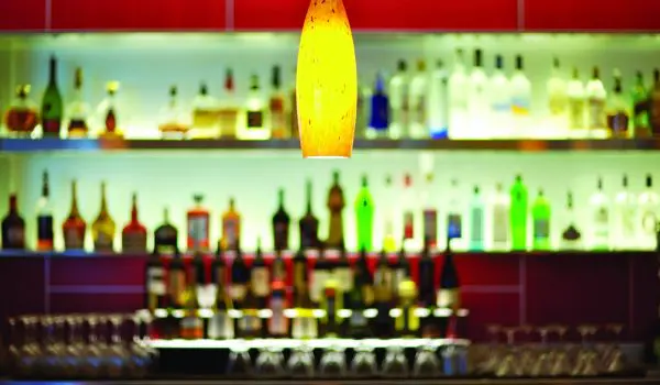 A warmly lit pendant lamp hangs above a bar counter lined with various bottles of alcohol against a red backdrop.