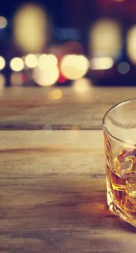 A glass of whiskey with ice on a wooden surface against a background of blurred lights.