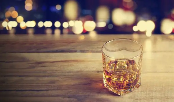 A glass of whiskey with ice on a wooden surface against a background of blurred lights.
