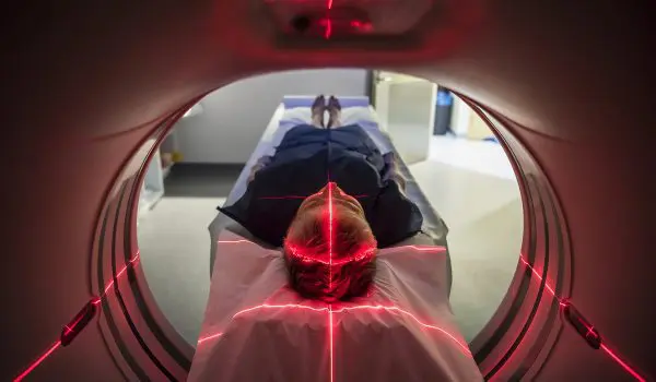 A person lies in a medical scanning device, possibly an MRI or CT machine, with red alignment lasers visible.