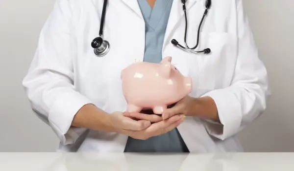 A healthcare professional wearing a lab coat with a stethoscope is holding a piggy bank.