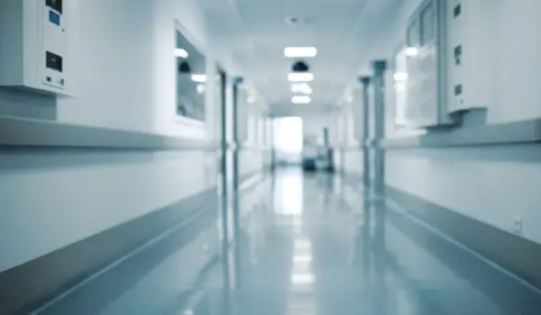 A blurry figure is visible at the end of a bright, clean hospital corridor with white walls and a reflective floor.