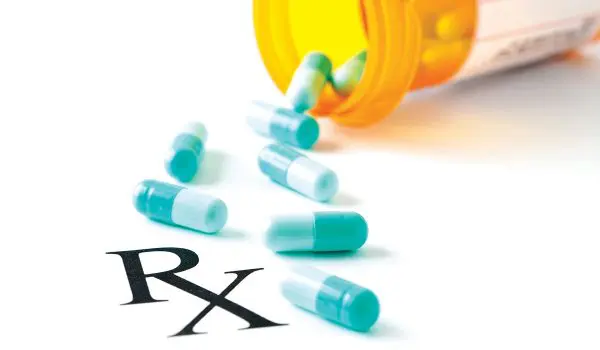 A prescription bottle lies on its side with blue and white capsules spilled out next to a bold "Rx" symbol on a white surface.