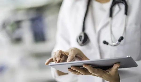 A healthcare professional wearing a white coat and stethoscope is using a digital tablet.