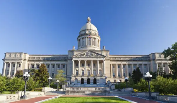 A grand neoclassical building with a large dome is basked in sunlight against a clear blue sky, surrounded by immaculate lawns and walking paths.