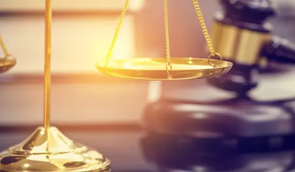 A set of golden scales is in focus in the foreground with a gavel and sound block blurred in the background, suggesting a legal or judicial setting.