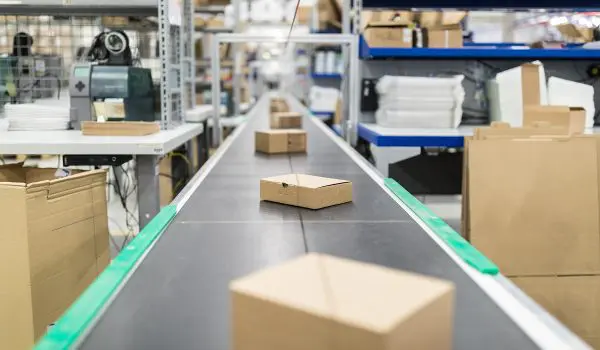 A conveyor belt in a warehouse carries cardboard boxes past shelves stocked with various items.