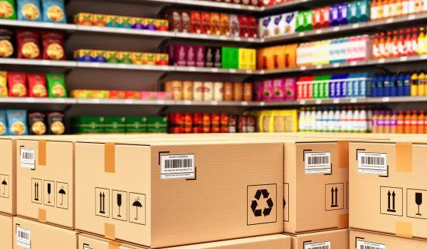 Cardboard boxes in the foreground with recycle symbols are stacked in front of colorful grocery shelves stocked with various products.