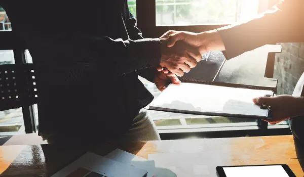 Two individuals are shaking hands over a table with documents and a digital tablet, suggesting a professional agreement or partnership.