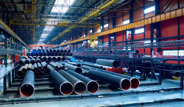 A collection of large industrial pipes is stored in a spacious factory with a high ceiling and overhead cranes.