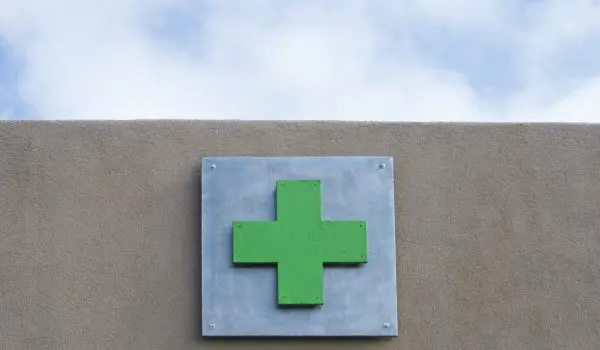A green cross sign, which often represents a pharmacy or medical services, is mounted on a wall under a blue sky with clouds.