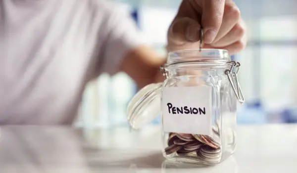 A hand is placing coins into a glass jar labeled "PENSION" on a table.