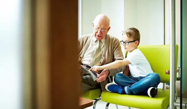 An elderly man and a young boy are sitting on a green bench, with the man showing something to the boy from a book or a tablet.