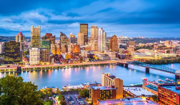 A panoramic twilight view of a city skyline with illuminated buildings reflected on a calm river.
